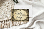 A square vintage-style RSVP card with ornate black filigree corners and cursive text reading 'KindlyRSVP' on a cream background, placed on top of a white envelope over a white draped fabric surface with a cream woven textile and a small green leaf visible.