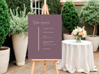 A wedding order of service sign on a wooden easel in an outdoor garden setting. The sign is a muted dusty purple with white text listing the order of service.