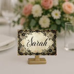 A personalized table place card on a small gold stand, featuring a vintage-style floral border and the name 'Sarah' in black script on a cream background, set against a blurred floral centerpiece.