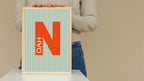 Person holding a framed poster with the letter 'N' and text on a beige background