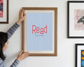 Person hanging a framed poster with 'Read' on a wall with other framed artworks.