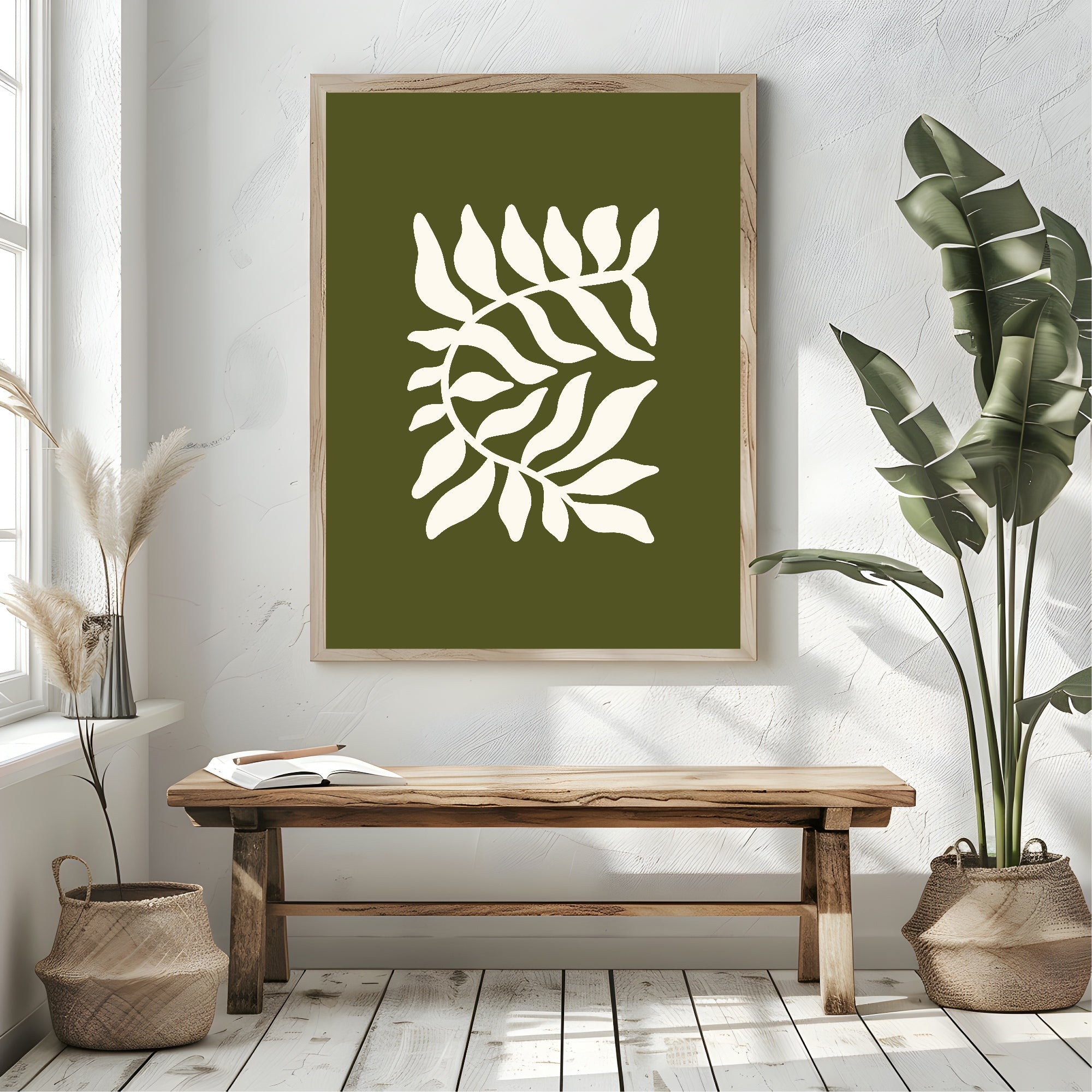 A framed botanical art print featuring a white leafy branch motif on an olive green background, styled in a bright room above a wooden bench with potted plants.