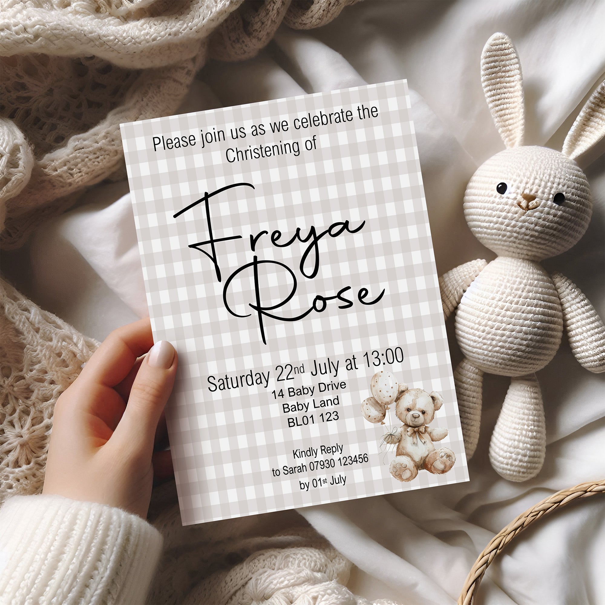 Invitation card for Freya Rose's christening with a teddy bear illustration, held by a hand on a soft fabric background.