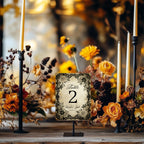 A small printed table number sign reading 'Table 2' stands on a small black easel in a styled event setting with candles, dried flowers, and orange roses on a wooden table.