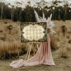 A rustic wedding seating plan sign with ornate black filigree borders and gold lettering, displayed on a wooden easel outdoors in a grassy field with pink draped fabric and pampas grass.
