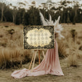 A rustic wedding seating plan sign with ornate black filigree borders and gold lettering, displayed on a wooden easel outdoors in a grassy field with pink draped fabric and pampas grass.