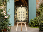 A wedding seating plan sign with an ornate black damask border on a cream background, displayed on a wooden easel in an outdoor garden setting with pink roses and light blue doors.