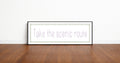 Framed print with 'Take the scenic route' text on a wooden floor and white wall background