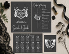 A styled flat-lay of a wedding invitation suite on a light beige background, surrounded by black lace, dried flowers, a black rose, and ribbon. The suite includes a main invitation, an order of the day panel, a seating chart, and a place card, all in a dark charcoal gray color with white text and skeleton hand heart illustrations.