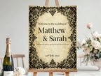 Wedding welcome sign with decorative frame on an easel, accompanied by a bottle of champagne and flowers.